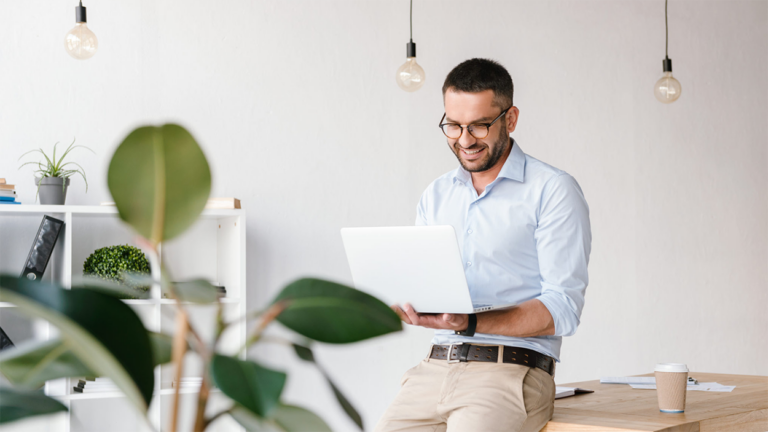 Male executive sitting on a desk looking at a laptop in an office setting