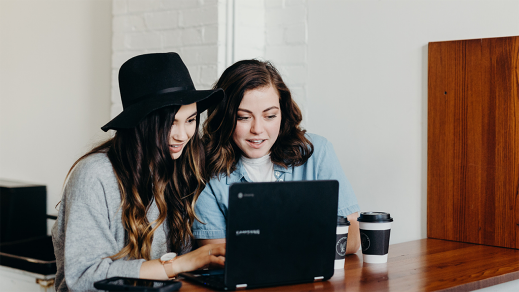 Two young women looking at a computer while sitting at a desk with coffee