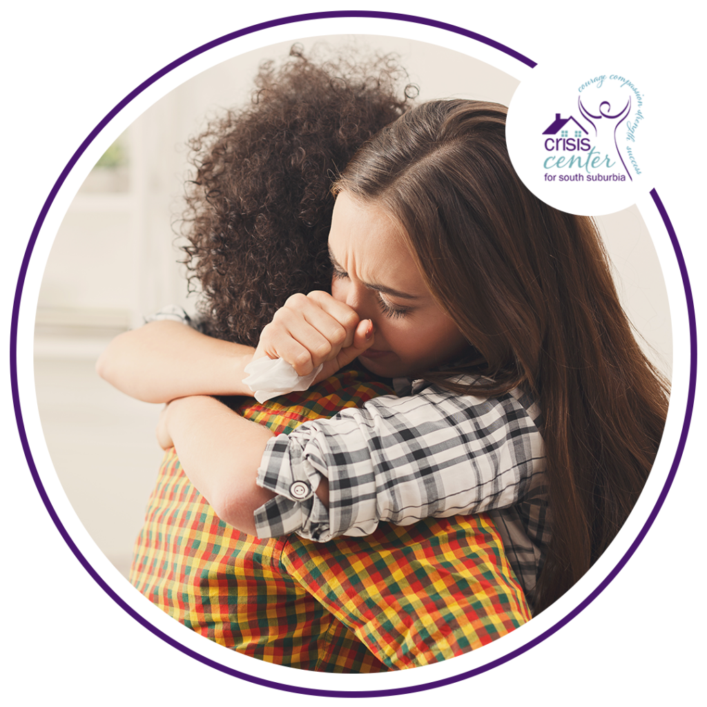 Image of a young woman hugging another person while holding a tissue with the logo of the Crisis Center for South Suburbia