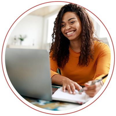 Image of a young woman wearing an orange shirt writing something down with a laptop in front of her