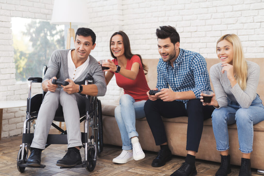 A group of friends are sitting down on a couch with one, in a wheelchair, holding playstation controllers and laughing together.