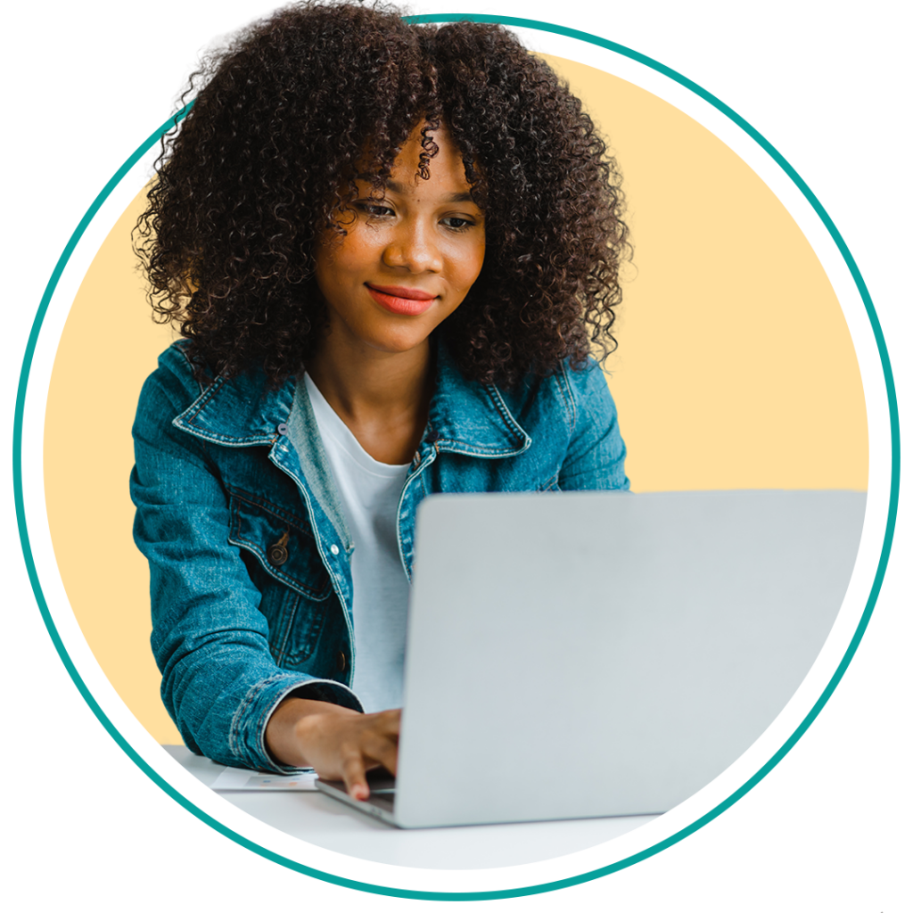 A young woman with curly hair working on a laptop. She is wearing a denim jacket over a white shirt and appears focused on her task