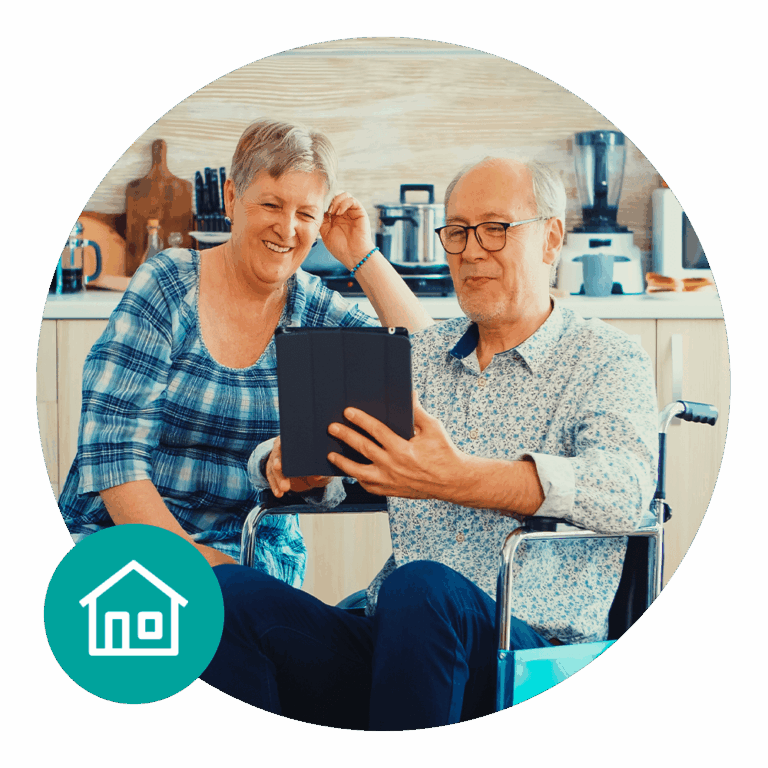 An older couple sitting together in a kitchen, smiling and looking at a tablet, with one person using a wheelchair and a small house icon in the corner.